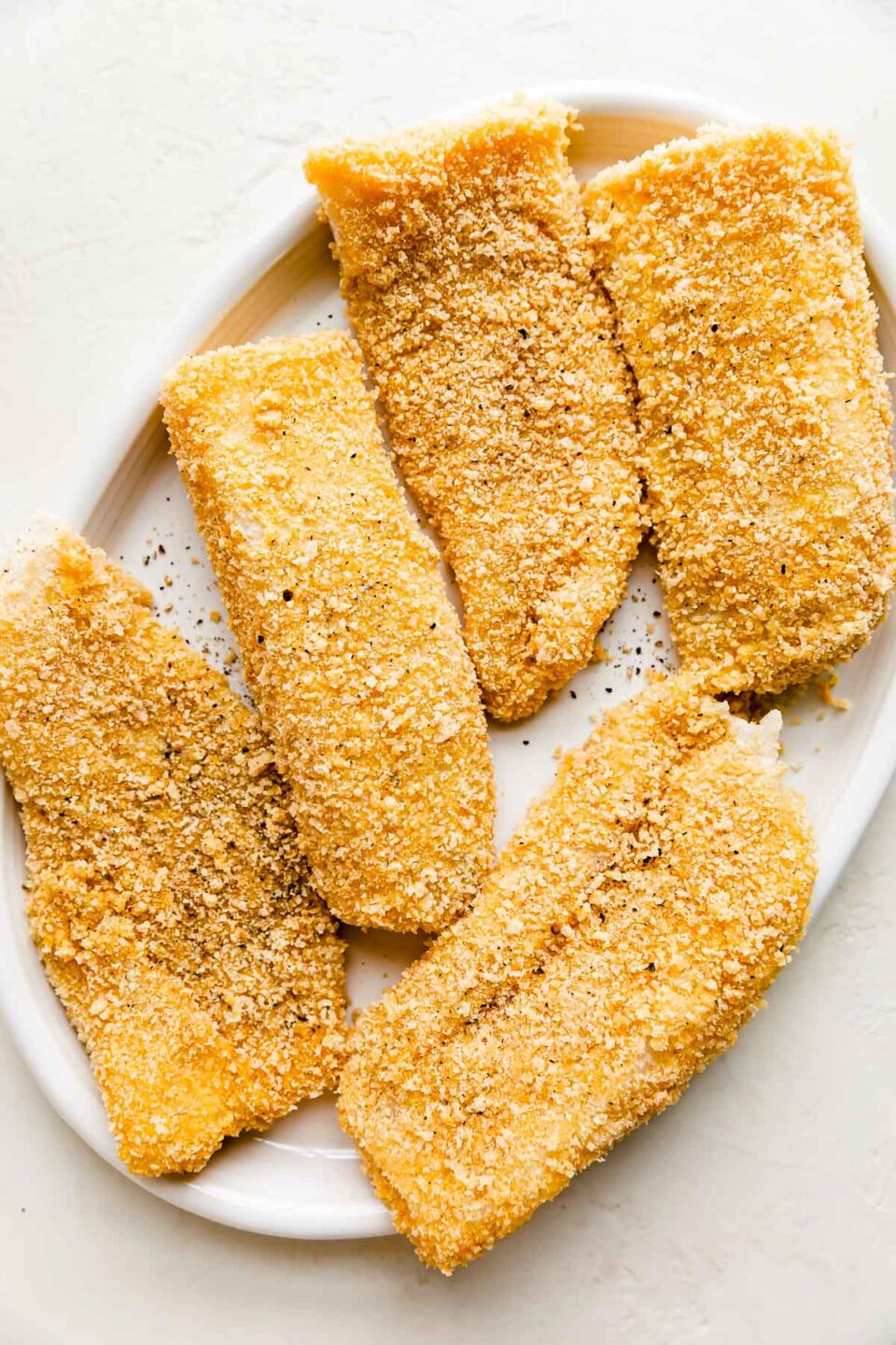 Five panko breaded cod fillets rest atop a white oval shaped serving platter before being shallow fried. The platter sits atop a creamy white textured surface.
