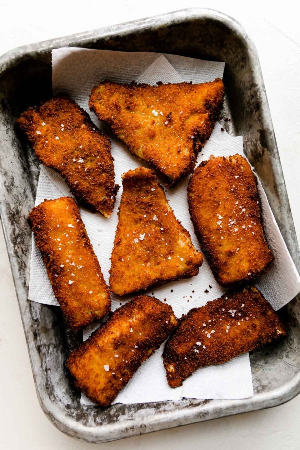 Seven pieces of fried cod rest atop a paper towel lined metal baking sheet. The baking sheet pan sits atop a creamy white textured surface and the fish has been garnished with flaky sea salt.