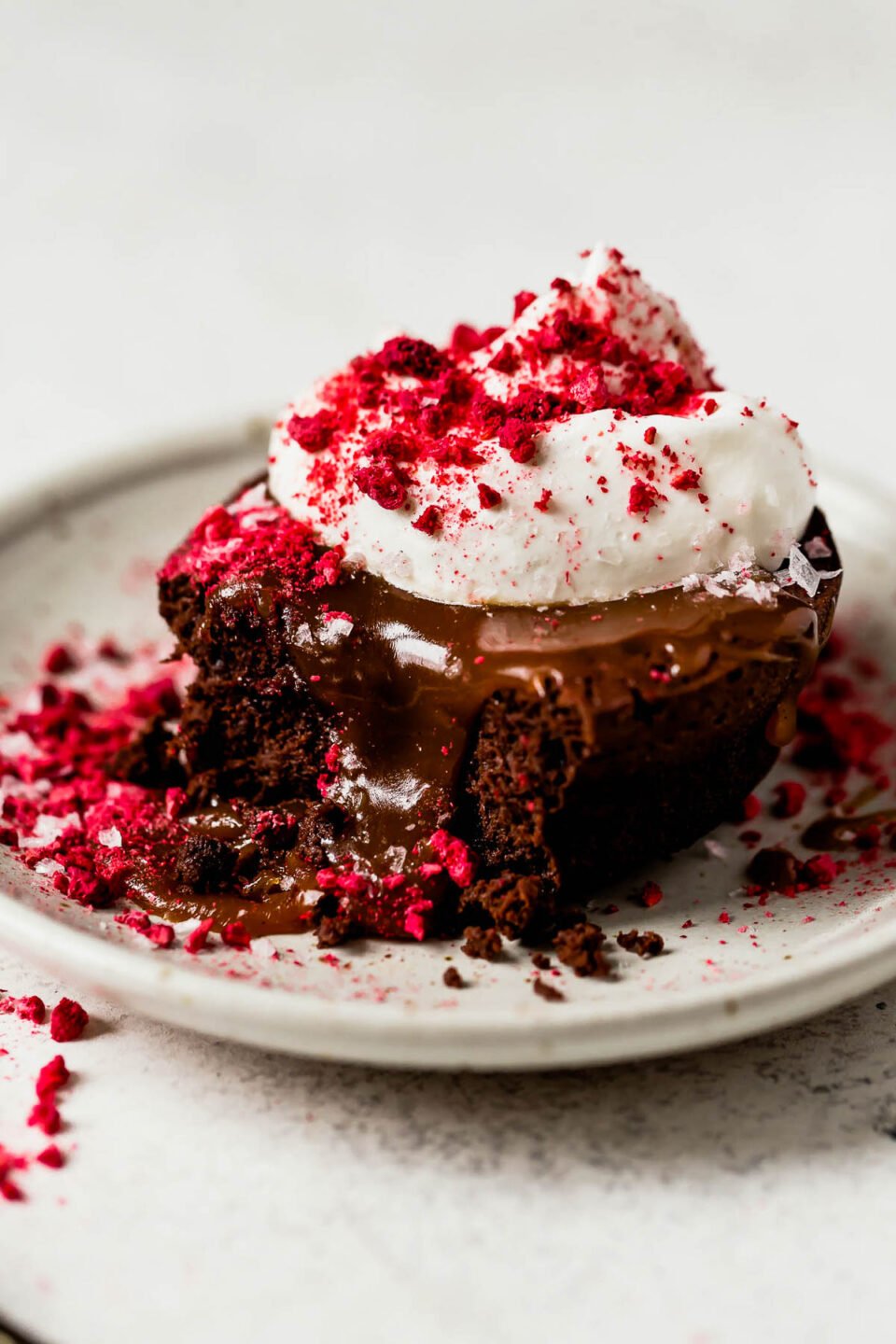 A side angle shot of a mini flourless chocolate cake that sits atop a light colored ceramic plate. The cake has been topped with caramel, whipped cream, crushed freeze-dried raspberries, and flaky sea salt. The plate sits atop a white textured surface.