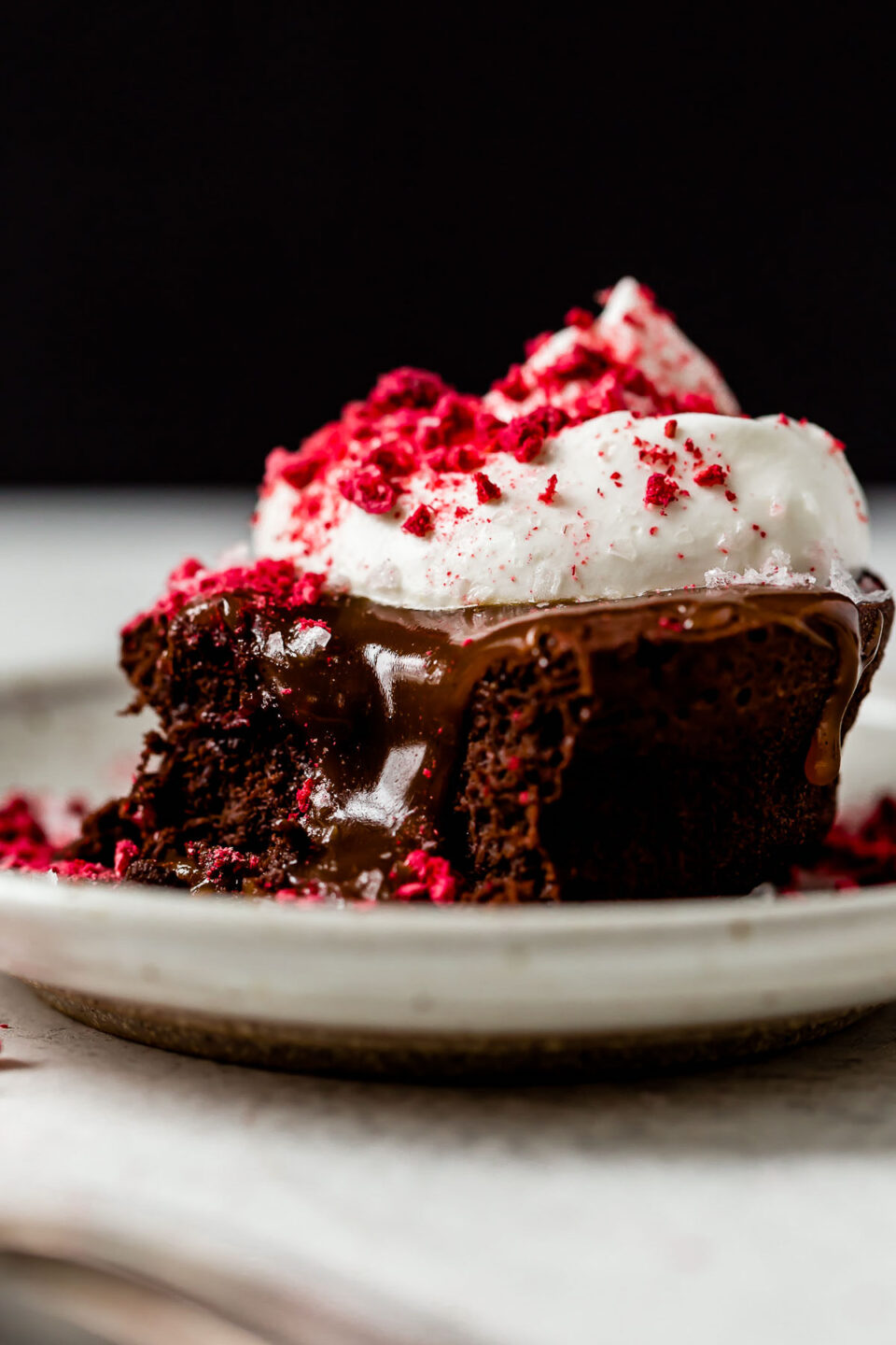 A mini flourless chocolate cake rests atop a white ceramic plate on a creamy white textured surface. The cake has been topped with caramel, whipped cream, crushed freeze-dried raspberries, and flaky sea salt.