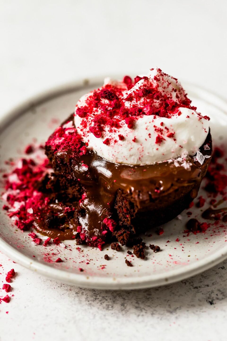 A slightly overhead and side angle shot of a mini flourless chocolate cake that sits atop a light colored ceramic plate. The cake has been topped with caramel, whipped cream, crushed freeze-dried raspberries, and flaky sea salt. The plate sits atop a white textured surface.