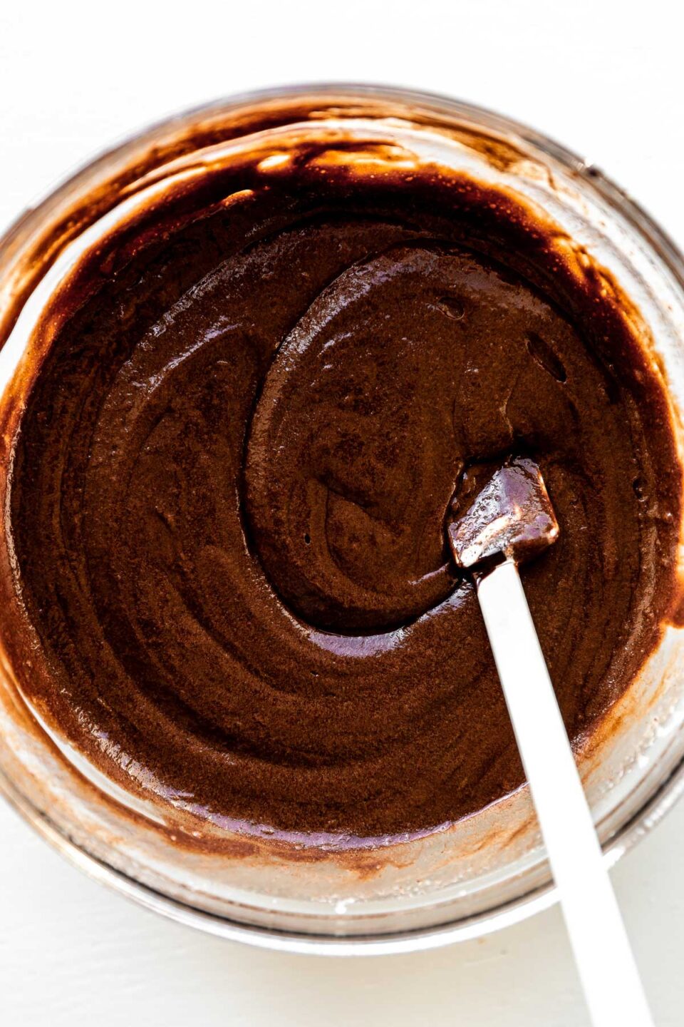 A tempered mixture of melted chocolate, eggs, sugar, and salt fill a large glass mixing bowl that sits atop a white textured surface. A small white spatula rests inside of the bowl.