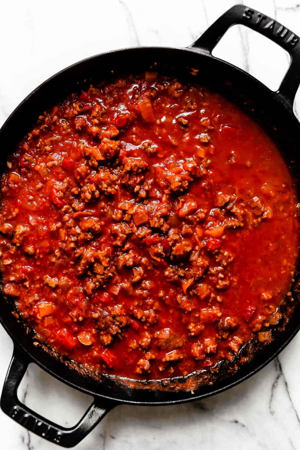 Italian sausage and peppers tomato sauce fills a large black Staub double handled skillet. The skillet sits atop a white and gray marble surface.