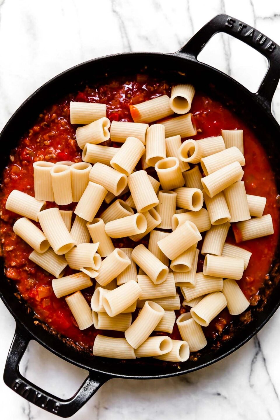 Rigatoni pasta is added to Italian sausage and peppers tomato sauce inside of a large black Staub double handled skillet. The skillet sits atop a white and gray marble surface.