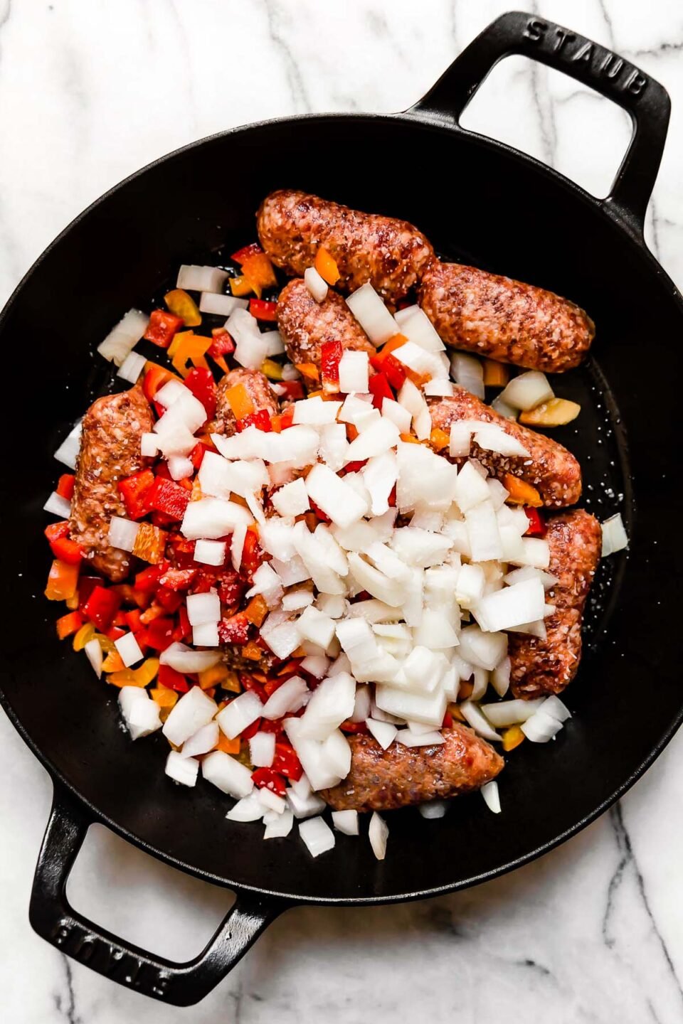 Italian sausage, diced bell pepper and onion inside of a large black Staub double handled skillet. The skillet sits atop a white and gray marble surface and the ingredients have been seasoned with kosher salt and black pepper.