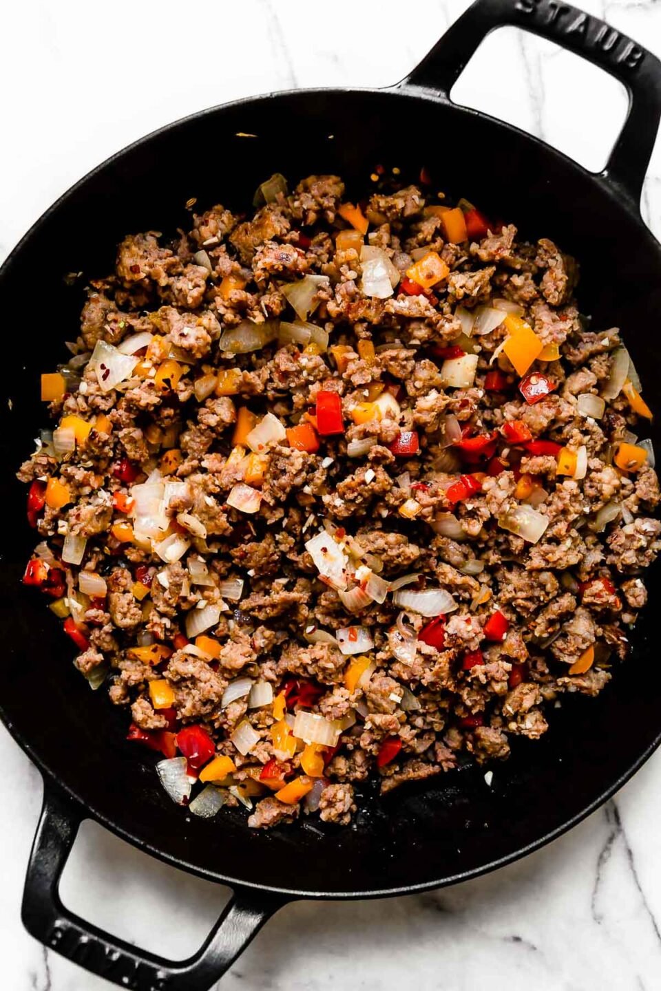 Crumbled and browned Italian sausage and softened bell pepper and onion fill a large black Staub double handled skillet. The skillet sits atop a white and gray marble surface.