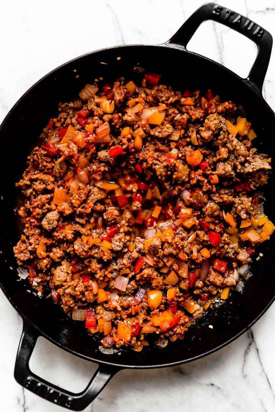 Crumbled and browned Italian sausage and softened bell pepper and onion mixed with tomato paste fill a large black Staub double handled skillet. The skillet sits atop a white and gray marble surface.