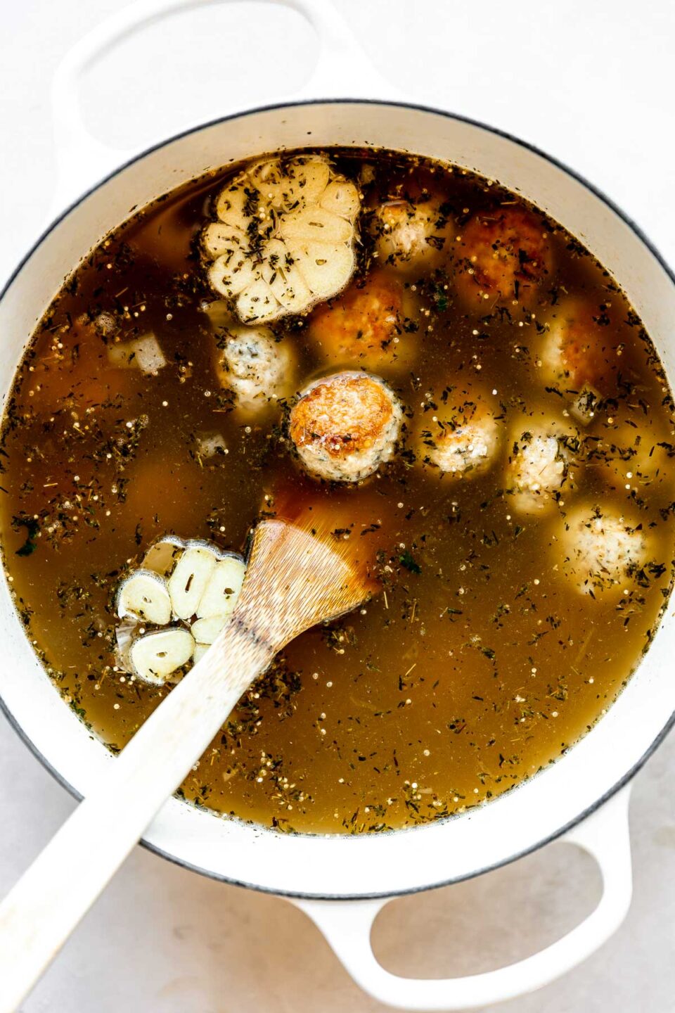 Italian wedding soup with turkey meatballs (or chicken meatballs) is built inside of a large white pot. The pot sits atop a creamy white textured surface.  A wooden turner rests inside of the pot for stirring.