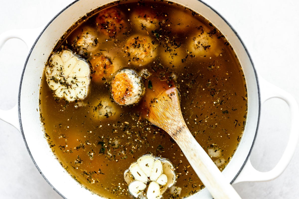 Italian wedding soup with turkey meatballs (or chicken meatballs) is built inside of a large white pot. The pot sits atop a creamy white textured surface. A wooden turner rests inside of the pot for stirring.