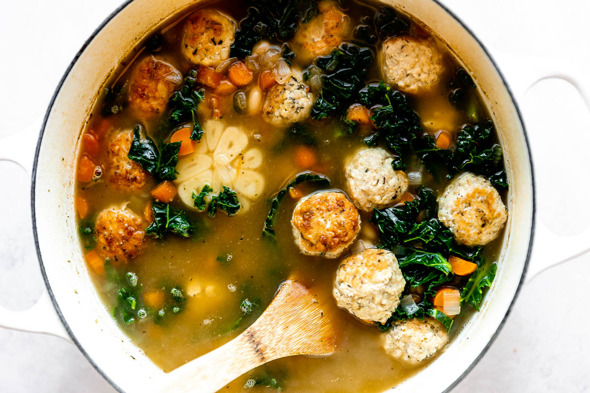 Italian wedding soup with meatballs simmers inside of a large white pot. The pot sits atop a creamy white textured surface. A wooden turner rests inside of the pot for stirring.