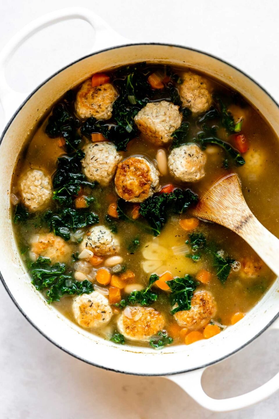 Italian wedding soup with meatballs simmers inside of a large white pot. The pot sits atop a creamy white textured surface.  A wooden turner rests inside of the pot for stirring.