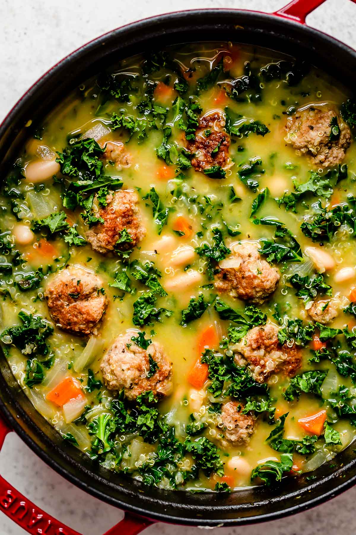 Italian wedding soup simmers inside of a large red pot. The pot sits atop a creamy white textured surface.