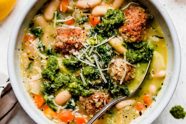 A close up and macro shot of Italian wedding soup with turkey meatballs served in a white bowl that sits atop a creamy white textured surface. The soup has been garnished with dollops of fresh pesto and shredded parmesan cheese. A spoon rests inside of the bowl.