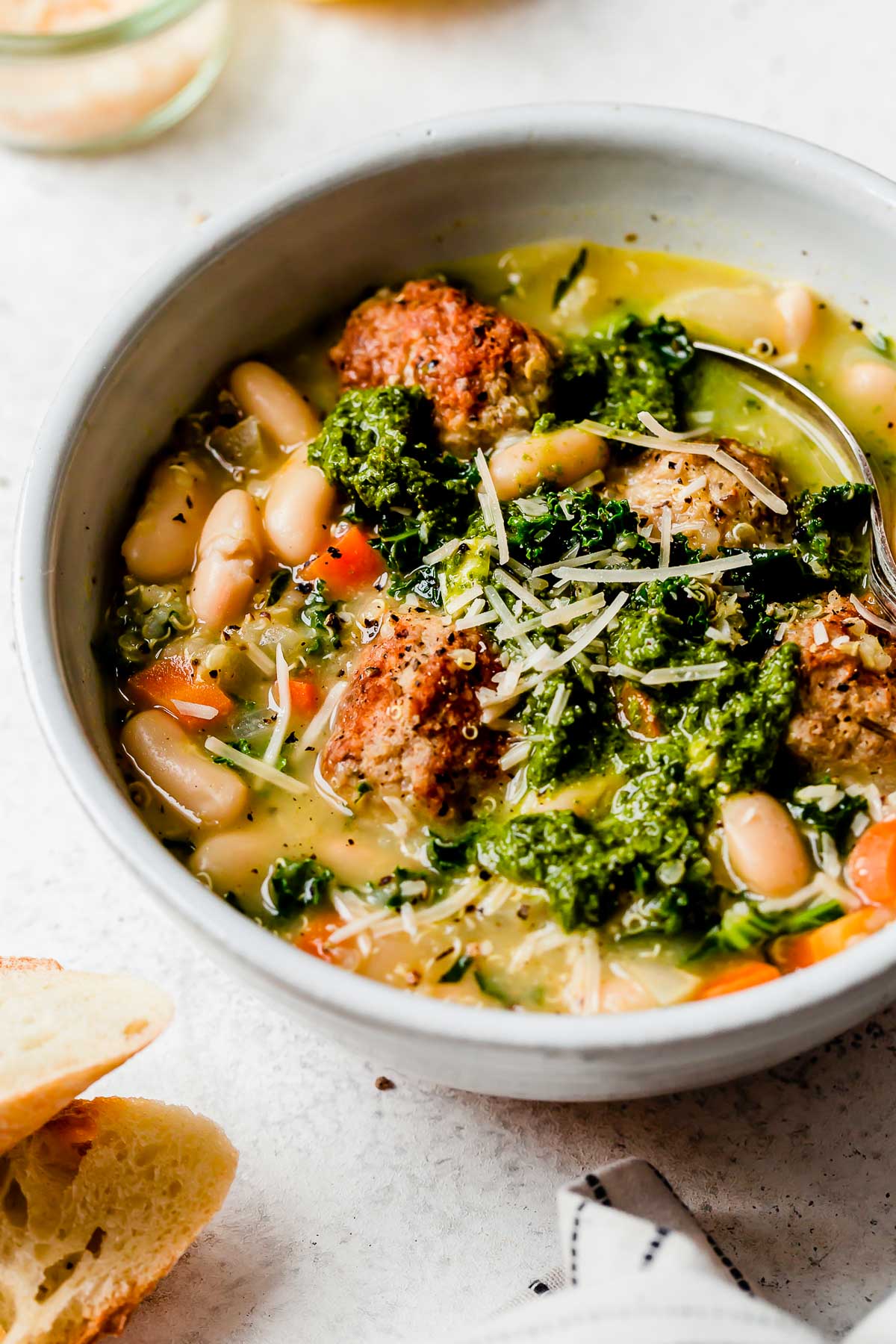 A side angle shot of Italian wedding soup with turkey meatballs served in a white bowl that sits atop a creamy white textured surface. The soup has been garnished with dollops of fresh pesto and shredded parmesan cheese. A spoon rests inside of the bowl and the bowl is surrounded by a white and black striped linen napkin, a few pieces of crusty bread, and a small glass jar filled with shredded parmesan cheese.