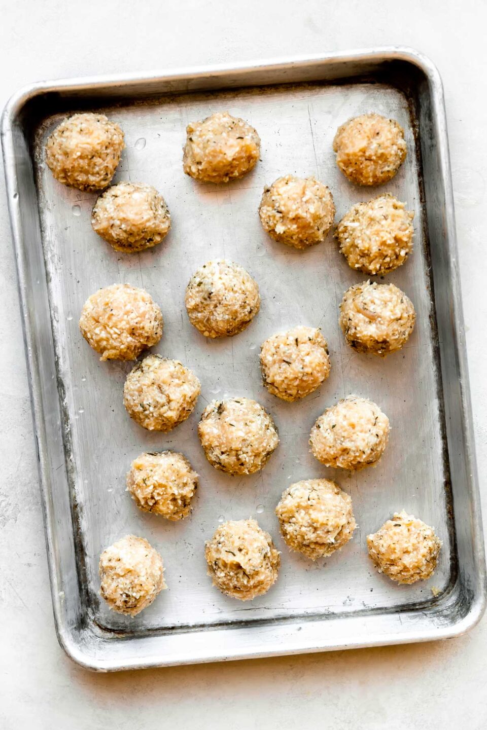 Eighteen formed chicken or turkey meatballs are arranged atop a metal baking sheet. The baking sheet sits atop a creamy white textured surface.