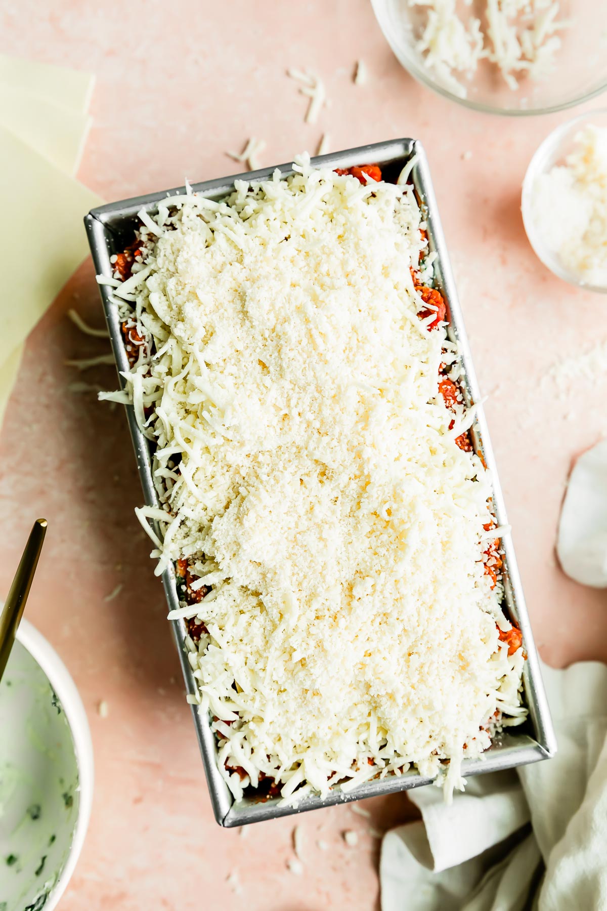 An assembled small batch lasagna is made inside of a loaf pan. The loaf pan sits atop a light pink surface and is surrounded by a bowl that was filled with spinach ricotta filling, a bowl of shredded mozzarella cheese, a small pinch bowl filled with grated parmesan cheese, lasagna sheets, and a light colored linen napkin.