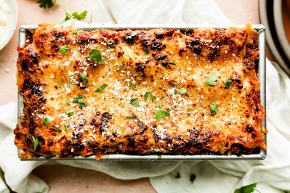 A close up shot of baked lasagna for two inside of a loaf pan that rests atop a light pink textured surface. A white linen napkin is placed underneath the loaf pan and the lasagna has been garnished with grated parmesan cheese and fresh chopped herbs.