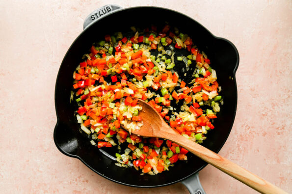 Diced bell peppers, onions, and celery are softened inside of a large gray Staub cast iron skillet. The skillet sits atop a light pink background and wooden spoon rests inside of the skillet.