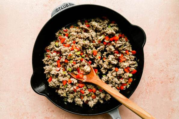 Diced and softened bell peppers, onions, and celery are combined with browned Italian sausage crumbles inside of a large gray Staub cast iron skillet. The skillet sits atop a light pink background and wooden spoon rests inside of the skillet.