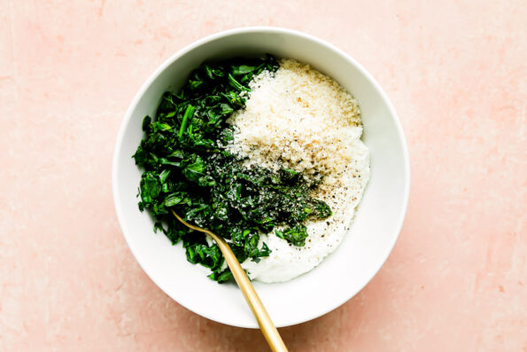 Wilted spinach, ricotta cheese, parmesan cheese, kosher salt & pepper fill a small white bowl that sits atop a light pink surface. A gold spoon rests inside of the bowl.