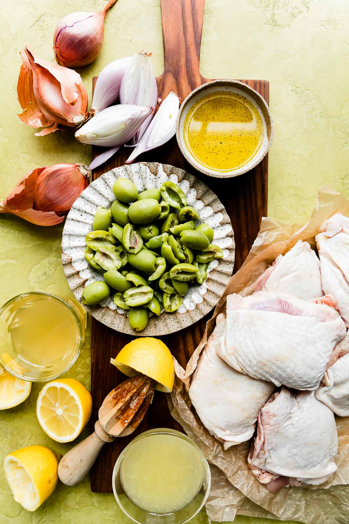 Olive chicken skillet ingredients laying on a yellow surface and wood cutting board: a dish of olive oil, a scalloped plate with green olives, chicken thighs, shallots, lemons (and lemon juice in a small bowl) & chicken stock in a small bowl.