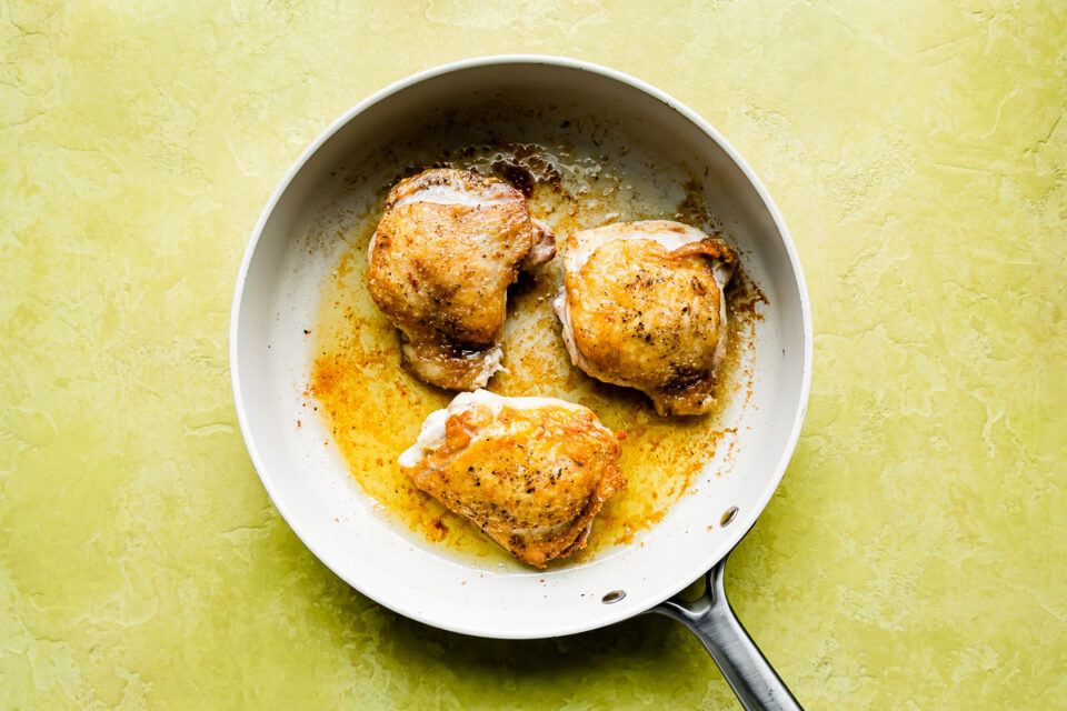 Three browned chicken thighs sit in a skillet atop a yellow textured surface.