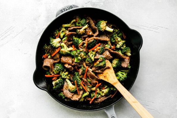 Beef broccoli stir fry fills a large gray Staub cast iron skillet that sits atop a creamy white textured surface. A wooden spoon rests inside of the pan for stirring.