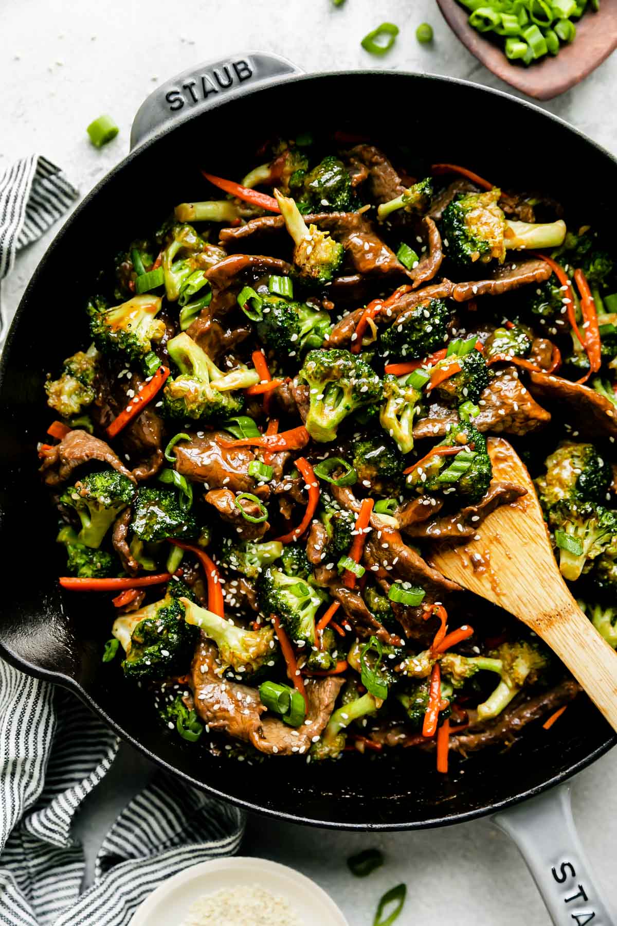 Broccoli and beef stir fry fills a large gray Staub cast iron skillet that sits atop a creamy white textured surface. A wooden spoon rests inside of the pan for stirring and the beef and broccoli stir fry is garnished with toasted sesame seeds and thinly sliced green onion. The pan is surrounded by a small wooden bowl filled with thinly sliced green onion, a blue and white striped linen napkin, and a small white plate filled with toasted sesame seeds.