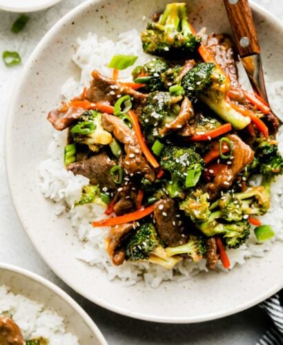 Beef and broccoli stir fry served over white rice fills a ceramic bowl. A fork with a wooden handle rests inside of the bowl and the bowl sits atop a creamy white textured surface. The bowl is surrounded by a small plateful of toasted sesame seeds, another bowl of beef broccoli stir fry served over rice, and a blue and white striped linen napkin.
