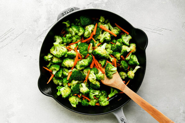 Broccoli florets and matchstick carrots stir fry inside of a large gray Staub cast iron skillet that sits atop a creamy white textured surface. A wooden spoon rests inside of the veggies for stirring.