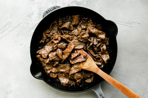 Seared and browned thinly sliced sirloin steak is combined with stir fry sauce inside of a large gray Staub cast iron skillet that sits atop a creamy white textured surface. A wooden spoon rests inside of the pan for stirring.
