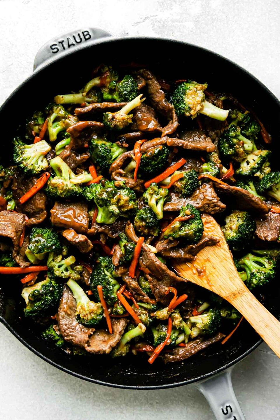 Beef and broccoli stir fry fills a large gray Staub cast iron skillet that sits atop a creamy white textured surface. A wooden spoon rests inside of the pan for stirring.