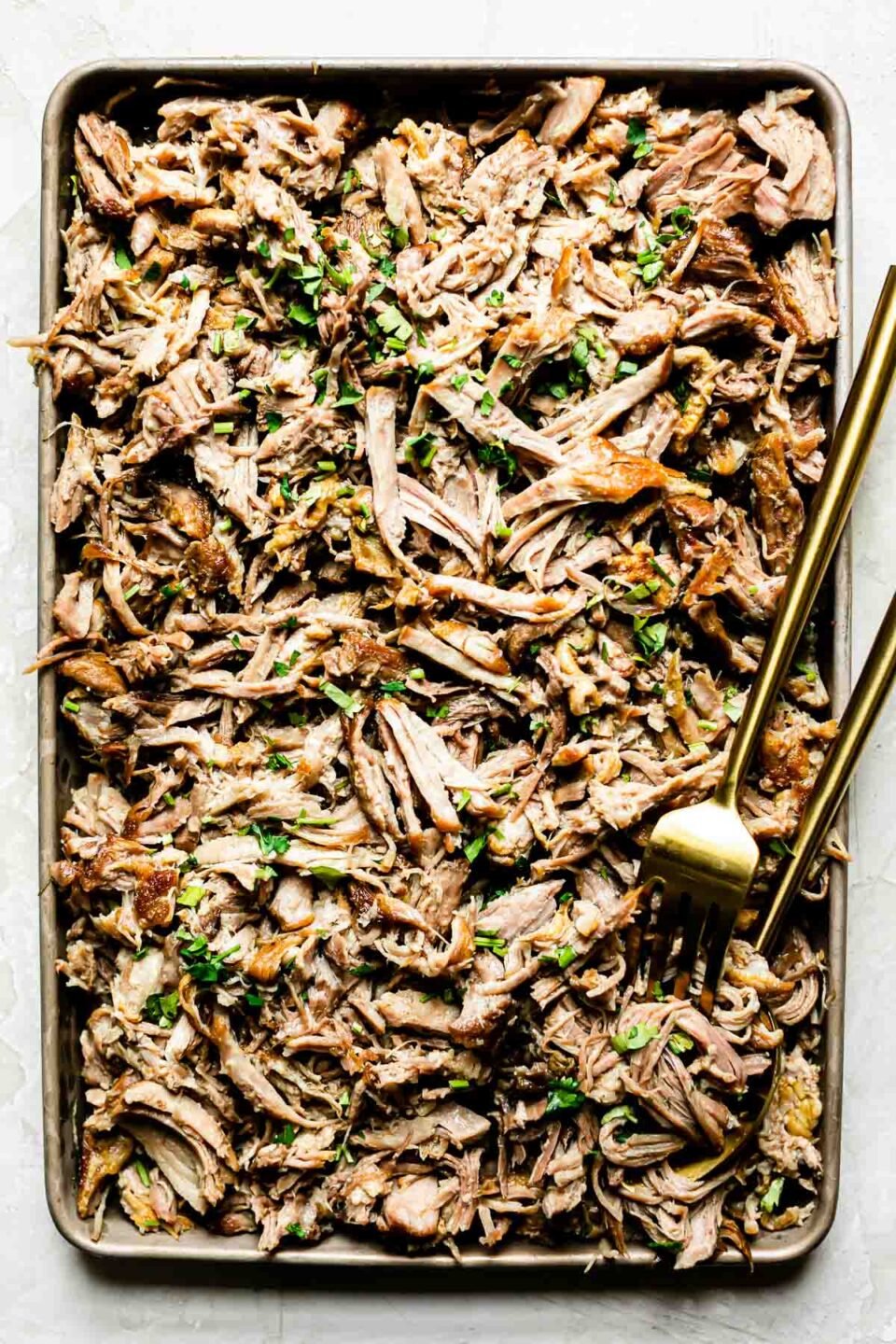 Shredded pork on a small baking sheet, topped with cilantro with a gold serving fork and spoon.