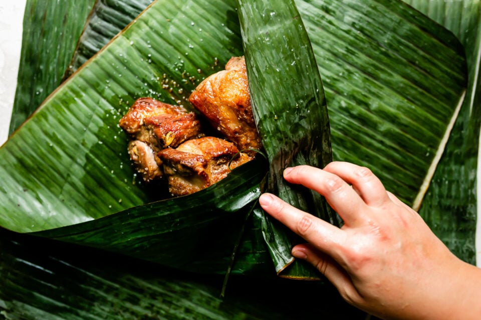 A woman's hands folds a banana leaf around browned pork.