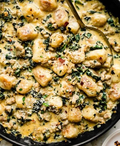 Sausage and gnocchi skillet fills a large black nonstick skillet that sits atop a creamy white textured surface. A gold spoon rests inside of the skillet and the skillet is surrounded by a small white plateful of crushed red pepper flakes and a white linen napkin with blue stripes.