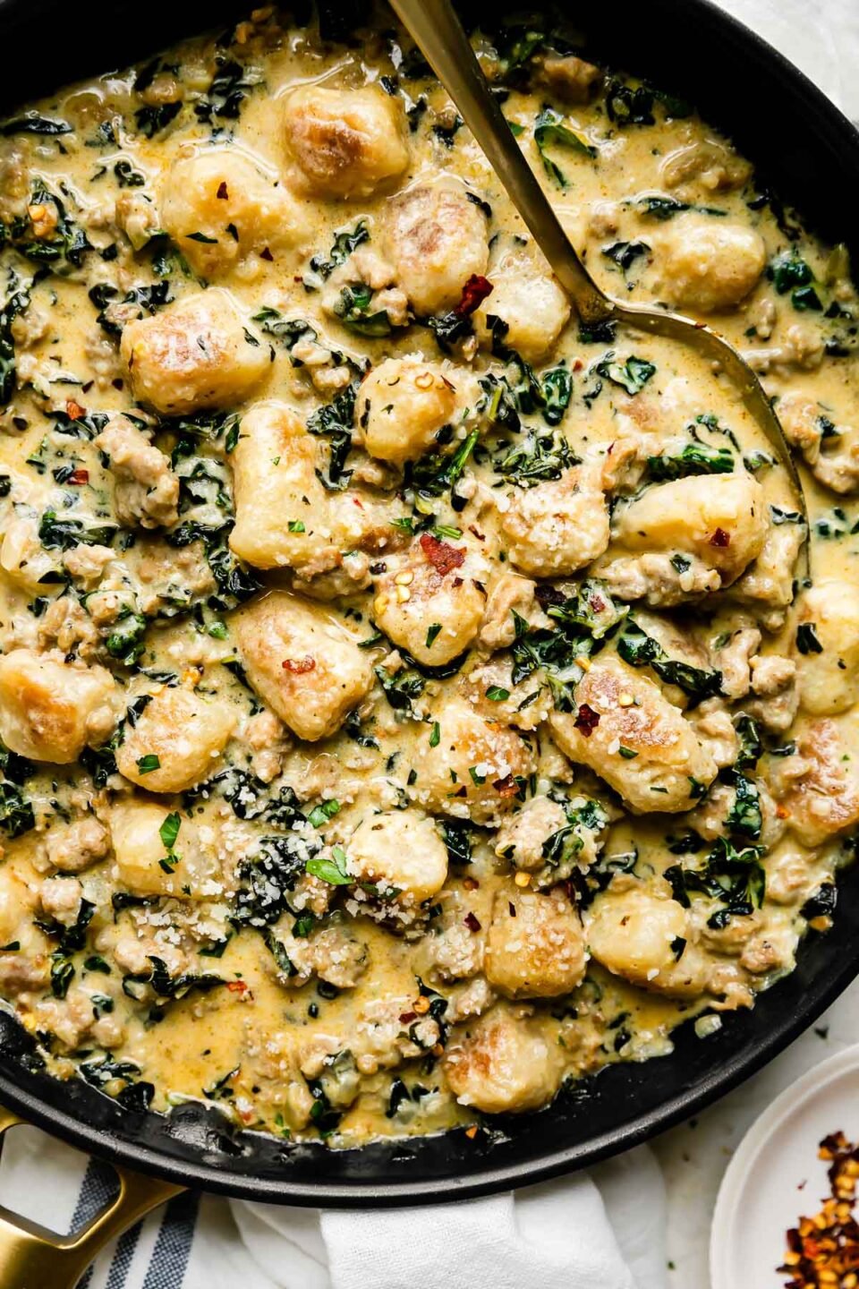 Sausage and gnocchi skillet fills a large black nonstick skillet that sits atop a creamy white textured surface. A gold spoon rests inside of the skillet and the skillet is surrounded by a small white plateful of crushed red pepper flakes and a white linen napkin with blue stripes.