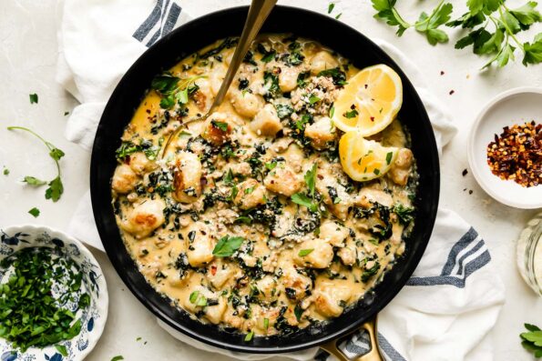 An overhead shot of gnocchi skillet inside of a large black nonstick skillet that sits atop a creamy white textured surface. A gold spoon rests inside of the skillet and the skillet is surrounded by a small white plateful of crushed red pepper flakes, loose fresh herbs, a small blue and white bowl of finely chopped fresh herbs, a glass of white wine, and a white linen napkin with blue stripes.
