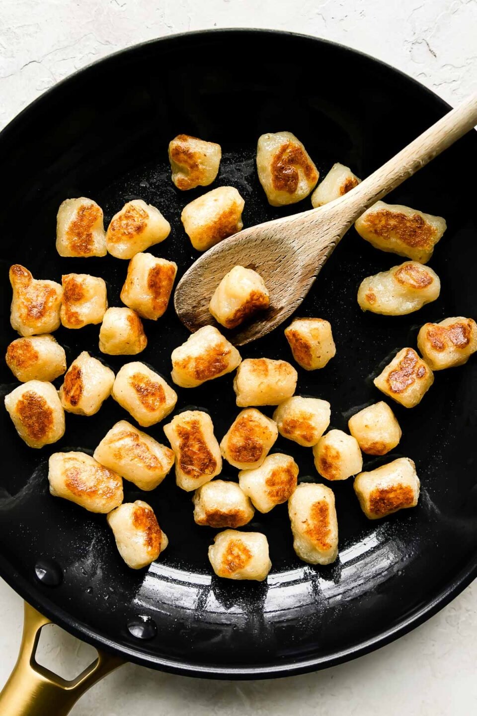 Cauliflower gnocchi fills a large black nonstick skillet that sits atop a creamy white textured surface. A wooden spoon rests inside of the skillet for stirring.