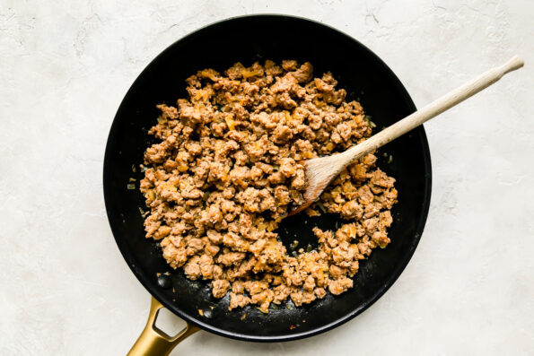 Browned mild or hot turkey Italian sausage fills a large black nonstick skillet that sits atop a creamy white textured surface. A wooden spoon rests inside of the skillet for stirring.