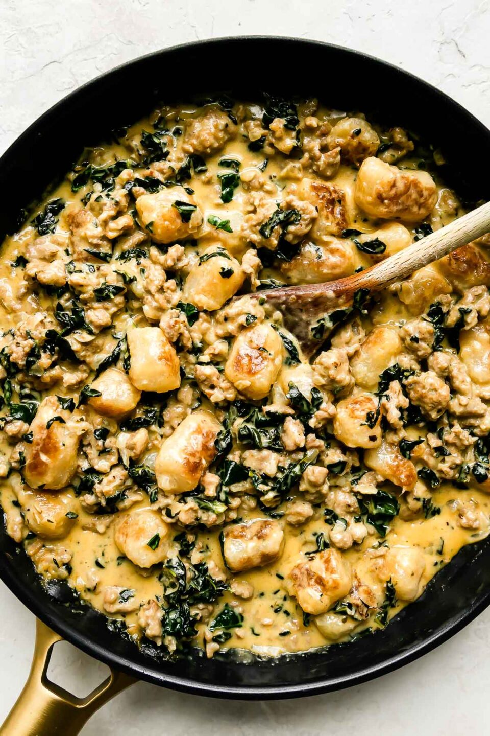 Cauliflower gnocchi skillet fills a large black nonstick skillet that sits atop a creamy white textured surface. A wooden spoon rests inside of the skillet for stirring.
