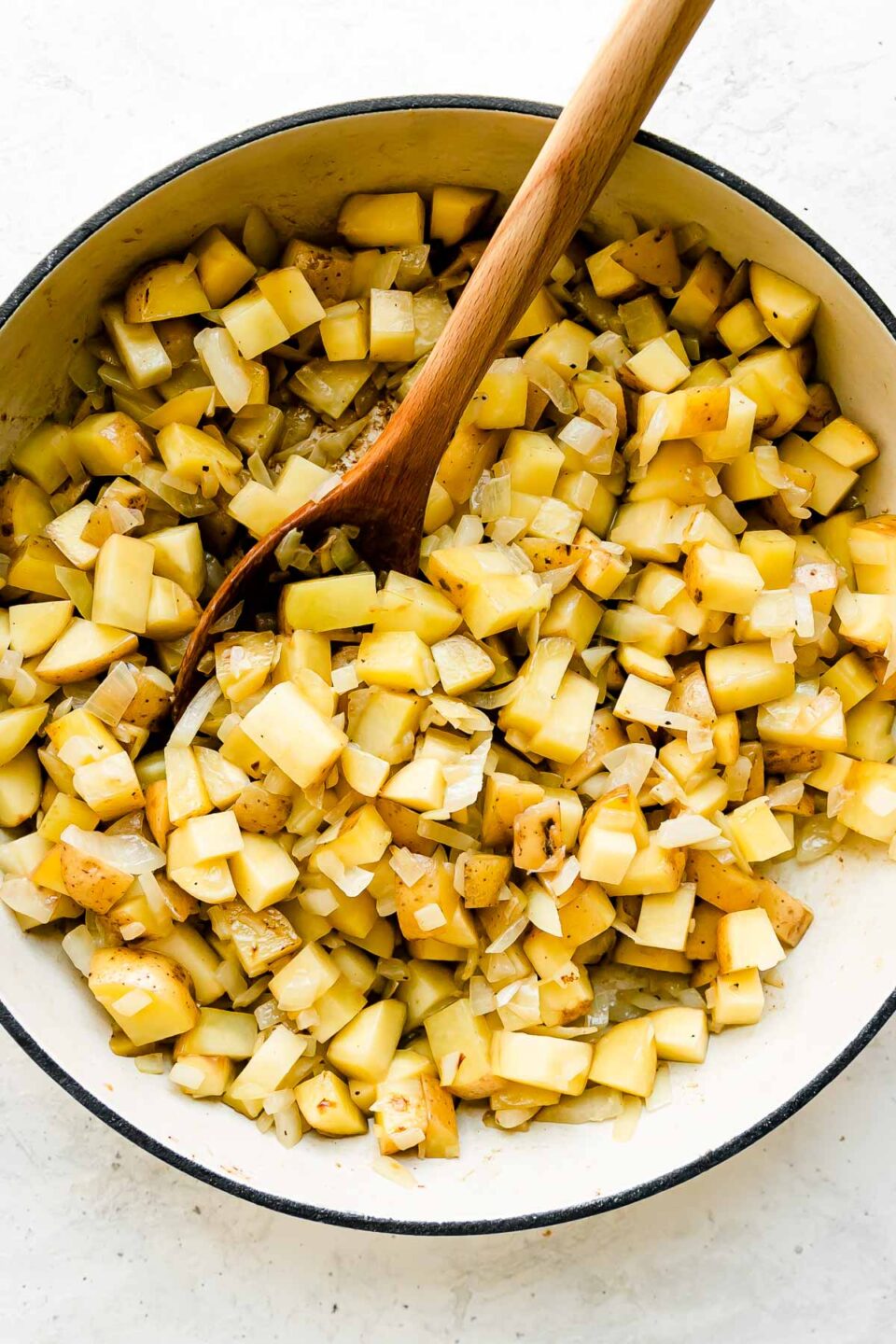 Diced Yukon gold potatoes and onion fill a large white double handled braising pan that sits atop a creamy white textured surface. A wooden spatula rests inside of the pan for stirring.