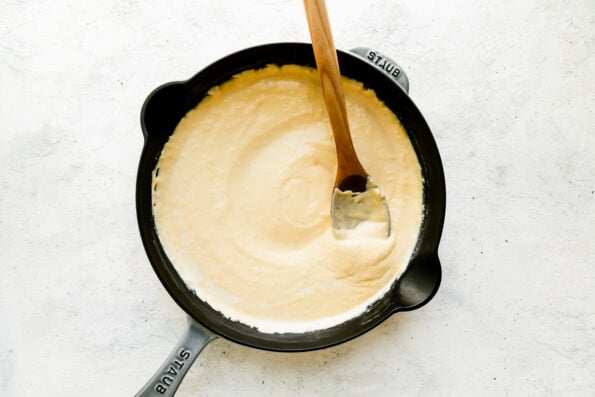 Homemade cheese sauce for cheesy ham and potato casserole fills a large gray Staub cast iron skillet that sits atop a creamy white textured surface. A wooden spoon rests inside of the skillet for stirring.