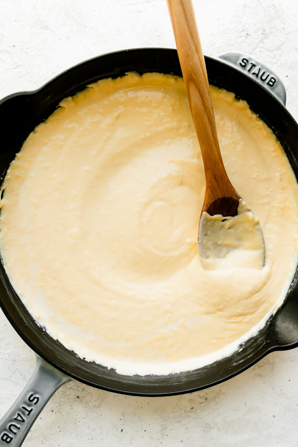 Homemade cheese sauce for cheesy ham and potato casserole fills a large gray Staub cast iron skillet that sits atop a creamy white textured surface. A wooden spoon rests inside of the skillet for stirring.