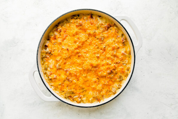 Baked ham and potato casserole fills a large white double handle braising pan that sits atop a creamy white textured surface.