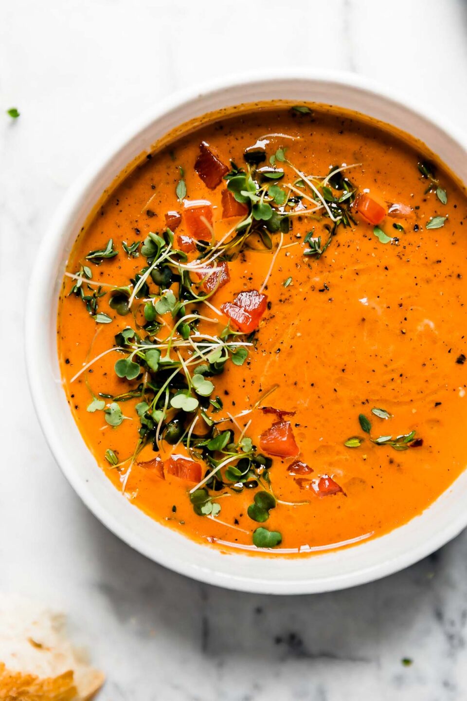 Creamy Roasted Red Pepper soup in a white ceramic bowl, topped with microgreens and cracked black pepper. The bowl is surrounded by some pieces of crusty bread. They are sitting atop a white marble surface.