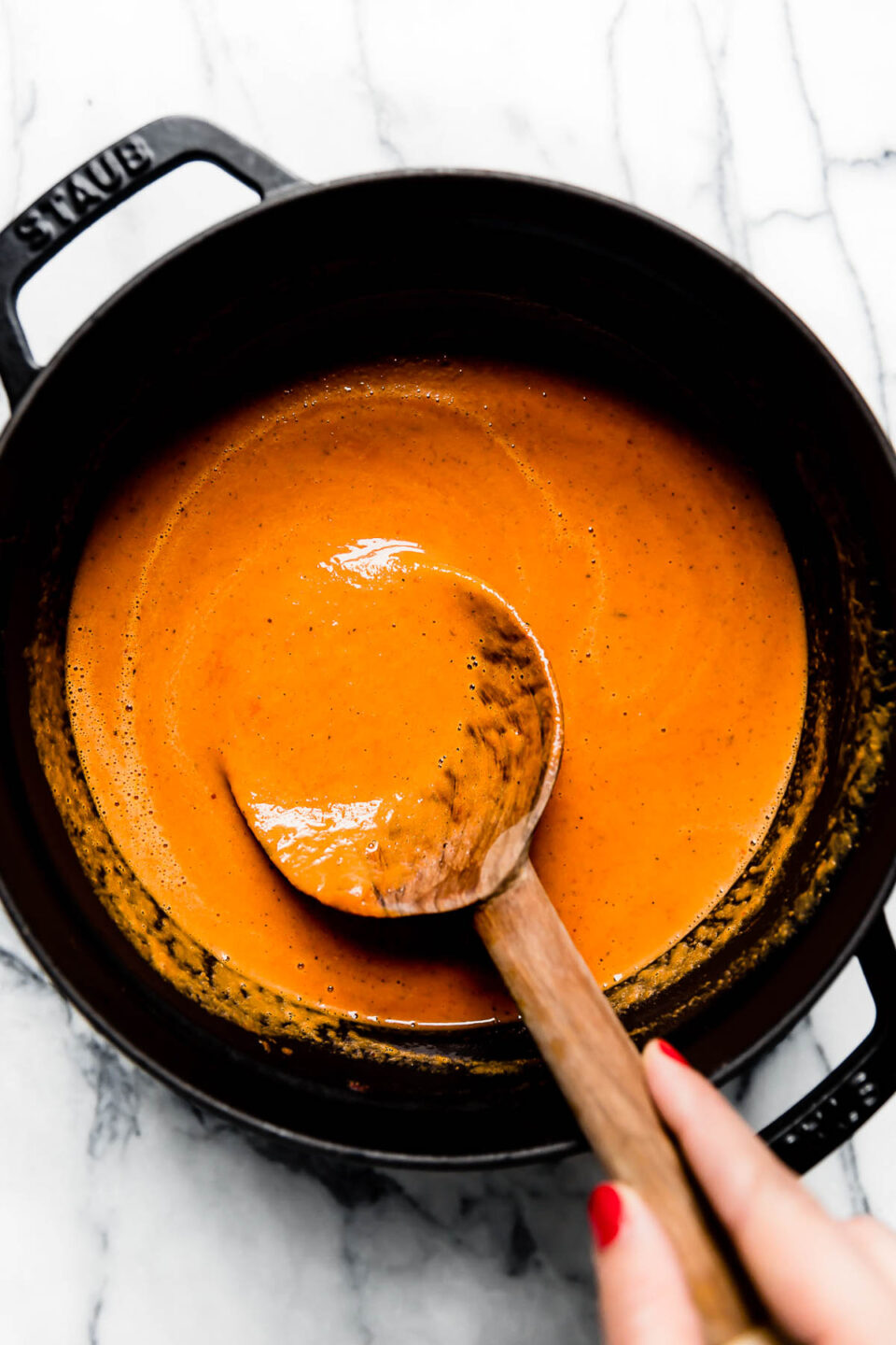 Creamy Roasted Red Pepper Soup, blended & topped with cracked black pepper, in a black Staub dutch oven. A woman's manicured hand is reaching into the Dutch oven with a wooden spoon to stir the soup.