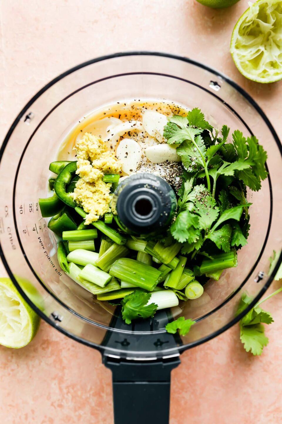 Green sauce ingredients fill a food processor bowl that sits atop a light pink textured surface: mayonnaise, buttermilk, lime juice, cilantro, jalapeno, fresh ginger, green onions, garlic, kosher salt, and ground black pepper. The food processor bowl is surrounded by discarded lime halves.