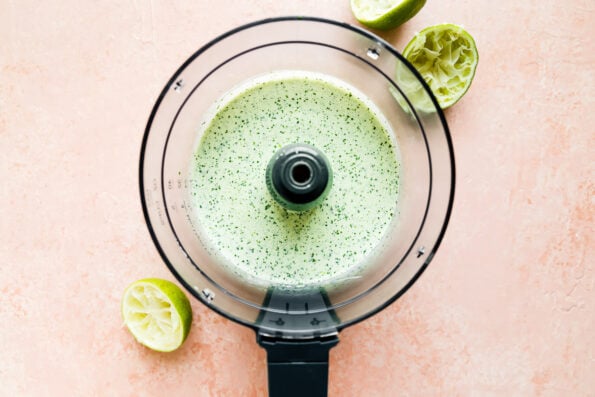 Prepared green sauce for salmon taco bowls fills a food processor bowl that sits atop a light pink textured surface. The food processor bowl is surrounded by discarded lime halves.