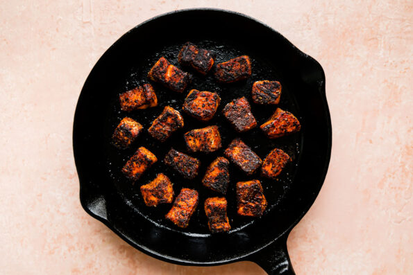 Seared cubed salmon fills a large black cast iron skillet that sits atop a light pink textured surface.