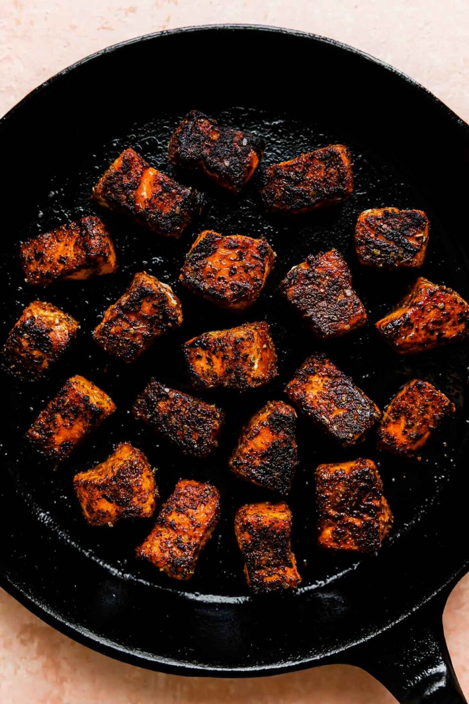 Seared cubed salmon fills a large black cast iron skillet that sits atop a light pink textured surface.